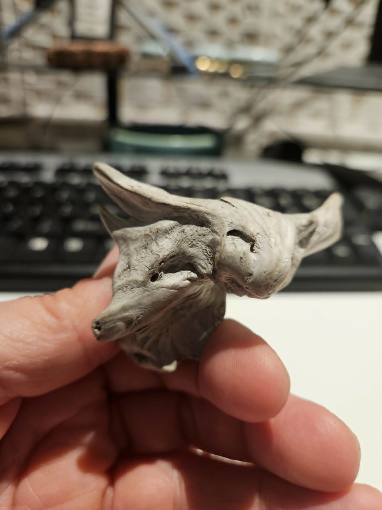 Small piece of salt-bleached driftwood held between thumb and finger; the grain and a dark knot suggest the face of a small canine, snout pointed left.